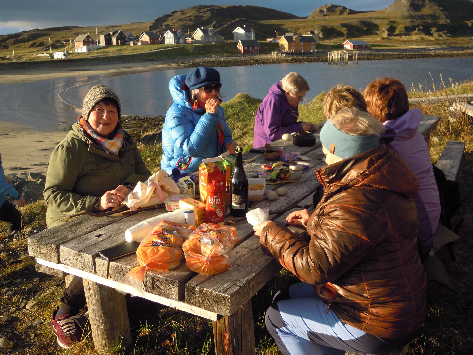 En gruppe mennesker sitter sammen på en benkl og spiser på tur ved vannet.