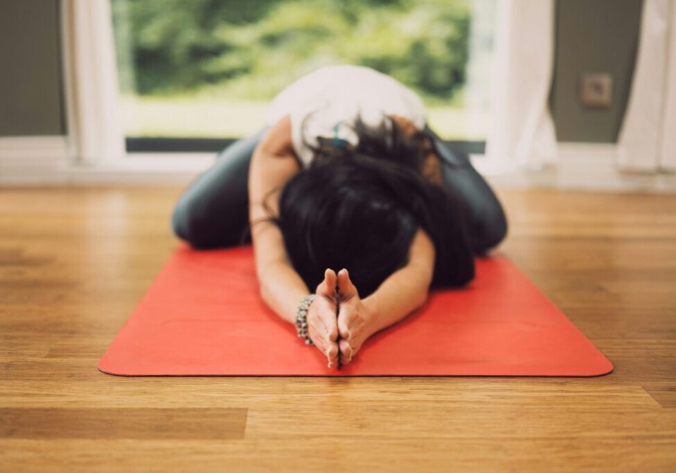 Kvinne gjør yogaøvelse liggende på gulvet, med armene over hodet vendt mot kamaera.