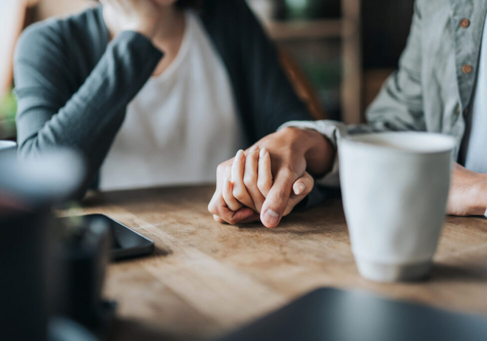 To personer sitter ved et bord, holder hender, viser omsorg. Vi ser kun hendene og en kaffekopp.